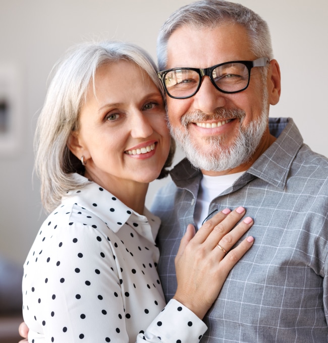 Smiling mature couple embracing indoors, woman in polka dot blouse, man wearing glasses and checkered shirt.