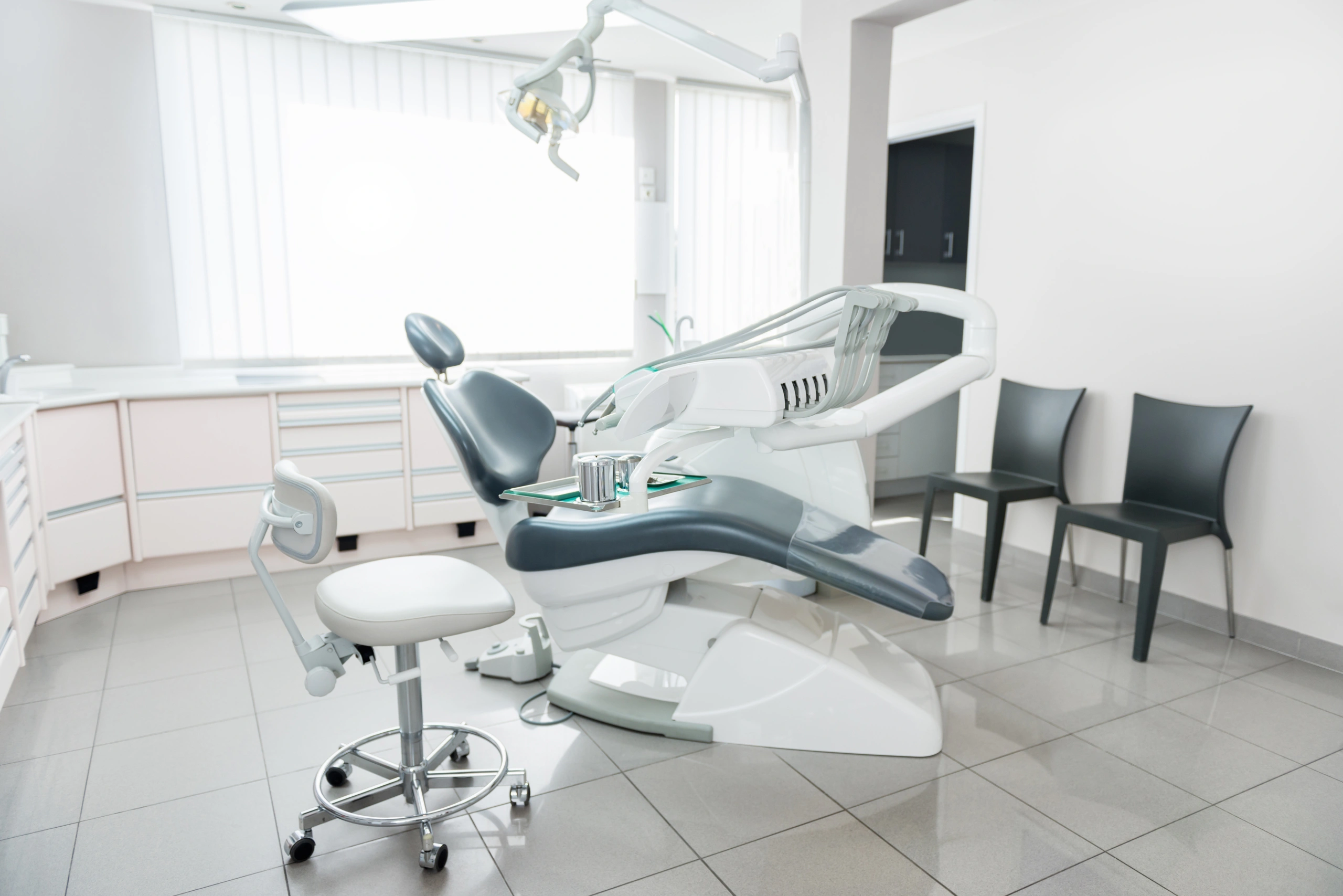 Modern dental operatory with blue chair, overhead lights, and patient stool in bright clinical setting.