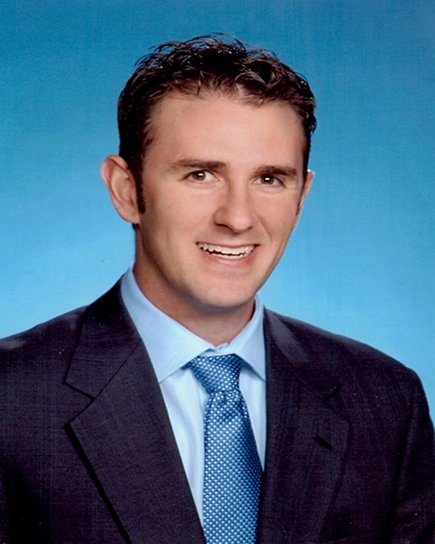Professional headshot of male dentist in navy suit and blue tie against blue background.