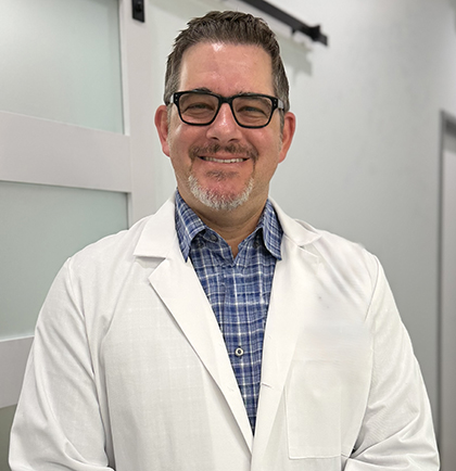 Male dentist wearing white lab coat and glasses smiles in clinical office setting.