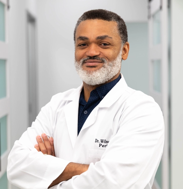 Male dentist in white coat with arms crossed in modern dental office setting.